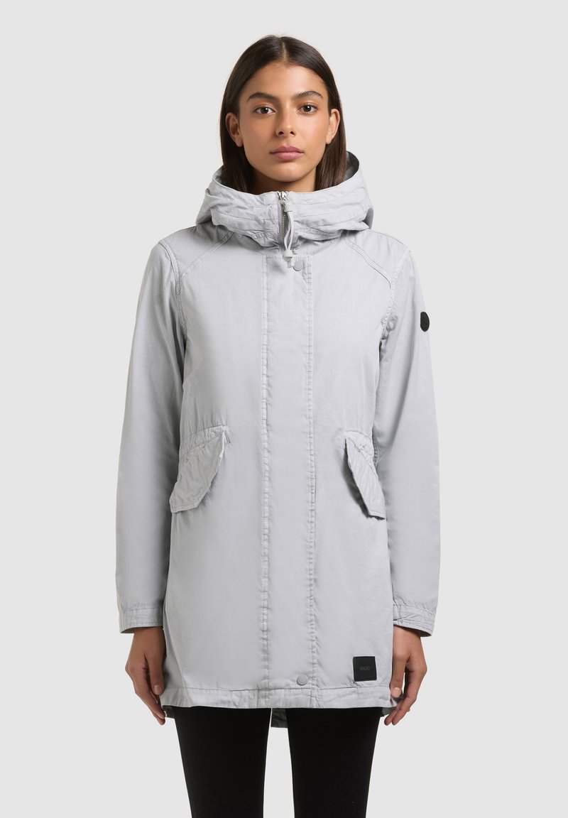 Femme de face portant un manteau à capuche gris clair avec des poches à rabat et un patch noir sur la manche gauche, sur un fond uni.