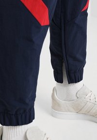 Pantalones deportivos de la marina con acentos rojos, puños elásticos y cremallera en el dobladillo. Combinados con zapatillas gris claro y calcetines blancos.