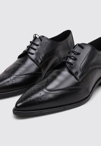 Zapatos de vestir de cuero negro con punta afilada, diseño de cordones y detalles de brogue en la parte superior. Textura suave con un brillo mínimo.