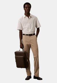 Camicetta a maniche corte a righe in bianco e azzurro chiaro, pantaloni beige, cintura nera, con una valigetta marrone, scarpe nere.