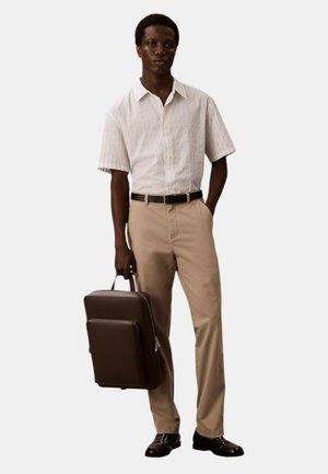 Camicetta a maniche corte a righe in bianco e azzurro chiaro, pantaloni beige, cintura nera, con una valigetta marrone, scarpe nere.