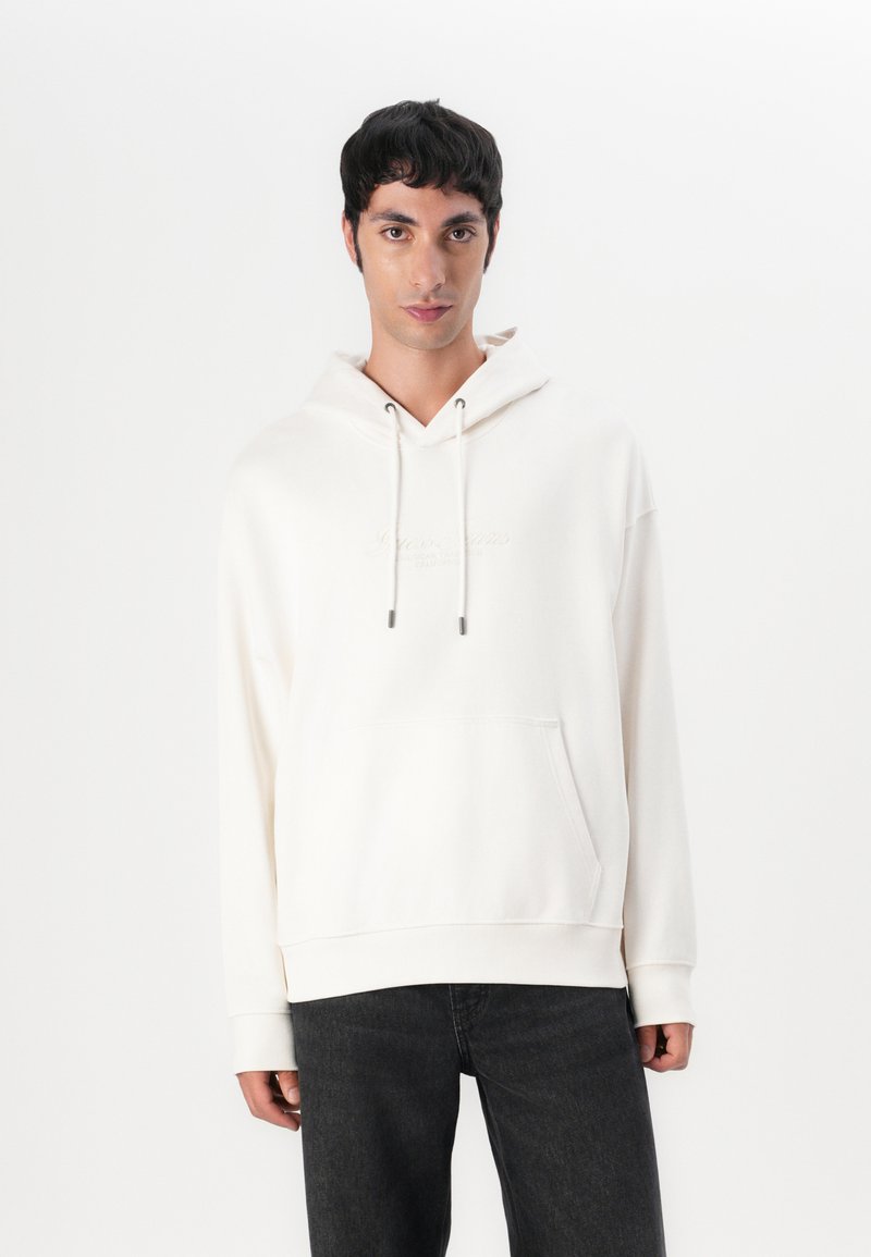 Weißer Kapuzenpullover mit einer Vorderstasche, ausgestattet mit einem dezenten geprägten Logo und einer Kordel, kombiniert mit dunklen Jeans. Weicher Stoff mit angenehmer Textur.