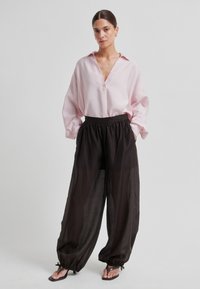 Světle růžová nadměrná košile na knoflíky s límečkem, spárovaná s černými, lehkými kalhotami se shromážděným pasem, zúženými nohavicemi a šňůrkami jako ozdobou.