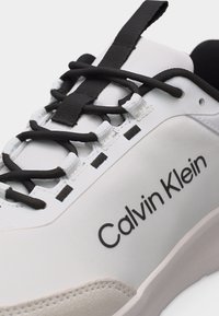 Λευκό και μαύρο αθλητικό παπούτσι Calvin Klein με μαύρα κορδόνια περασμένα από λευκές υφασμάτινες τρυπούλες και εμφανές λογότυπο της μάρκας στο πλάι.