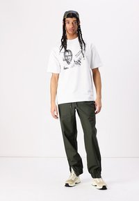 White graphic t-shirt with black print; loose fit. Green drawstring pants with elastic cuffs. Beige sneakers. Baseball cap with pattern.