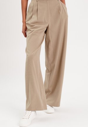 Pantalons beige à jambes larges en tissu doux avec des plis à la taille, associés à des baskets blanches. Pas de motifs ni d'accents visibles.