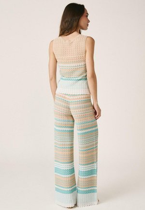 Mujer de pie de perfil, vestida con top de punto sin mangas y pantalones anchos con rayas en zigzag horizontales en beige, blanco, verde azulado y azul claro.