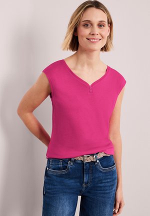 Mujer con cabello rubio corto, vestida con una blusa sin mangas de color rosa brillante y jeans azules, sonriendo con una mano detrás de la espalda.