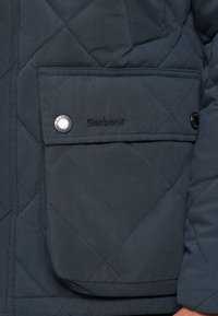 Σκούρο μπλε καπιτονέ μπουφάν με μπροστινή τσέπη· διαθέτει κουμπί τύπου snap και κεντημένο όνομα μάρκας "Barbour" σε μαύρο χρώμα. Ομαλή υφή, μινιμαλιστικός σχεδιασμός.