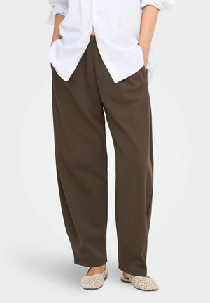 Personne portant un pantalon marron ample, une chemise blanche avec les manches retroussées, les mains dans les poches et des chaussures beiges tissées à enfiler.