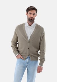Olivgrüner Cardigan mit V-Ausschnitt, knöpfigem Frontverschluss und strukturiertem Stoff, kombiniert mit einem hellen karierten Hemd und hellen Jeans.