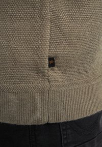 Pulover tricotat într-un verde oliv șters, cu o țesătură texturată, tiv ribotat și un mic etichetă neagră cu un logo portocalie pe cusătura laterală.