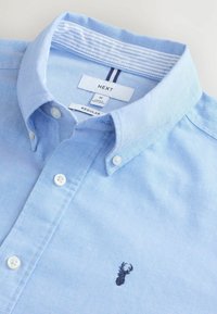 Chemise bleu clair à manches longues avec une finition texturée, ornée d'un logo de cerf brodé en noir et d'un col interne à rayures contrastées.