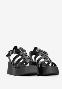 Sandalias de plataforma negras con tiras, hechas de material sintético; cuentan con una suela de cuña gruesa y una correa ajustable en el tobillo.