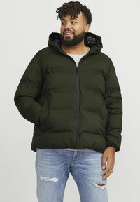 Chaqueta acolchada de color verde oliva con cremallera en el frente y capucha. Presenta un diseño acolchado, dos bolsillos laterales y forro negro. Lleva puesta una camiseta gris clara.