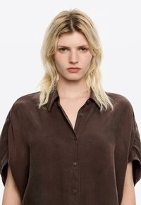 Brown, short-sleeved button-up shirt made of soft fabric. Features a classic collar and a relaxed fit, with a smooth, matte texture.
