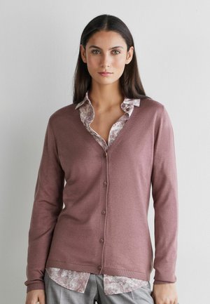 Vrouw met lang donker haar, gekleed in een mauvekleurig vest met knopen over een blouse met patroon en kraag, en grijze broek, staand tegen een lichte muur.