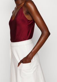 Bordeaux satijnen camisole met V-hals, gecombineerd met een witte schortachtige rok met een vo pocket en contrasterende stikdetails.