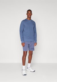 Sweatshirt azul com gola redonda e punhos canelados, combinado com calções cinzentos com letras grandes impressas. Sapatos e meias desportivas brancas.