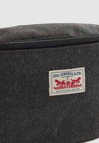 Dunkelgrauer, strukturierten Stoffbeutel mit schwarzem Reißverschluss und einem weißen Label von Levi Strauss & Co., das zwei rote Pferde zeigt, die Jeans ziehen.