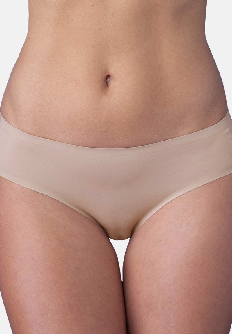 Środkowa część ciała kobiety pokazująca brzuch i górne partie ud w bezszwowej, cielistej bieliźnie na jednolitym tle.