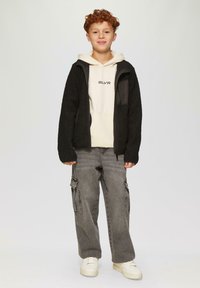 Čierna, textúrovaná fleecová bunda so zipsom, nosená cez smotanovú mikinu s potlačou "SLVR". Šedé cargo nohavice a biele tenisky dotvárajú outfit.