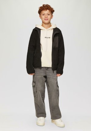 Zwarte, gestructureerde fleece jas met ritssluiting, gedragen over een crèmekleurige hoodie met "SLVR" print. Grijze cargobroek en witte sneakers maken de outfit compleet.