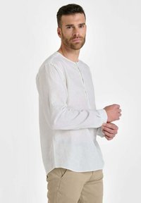 Man met kort donker haar en baard past de manchet van een wit overhemd met lange mouwen aan, gekleed in beige broek, tegen een effen lichte achtergrond.