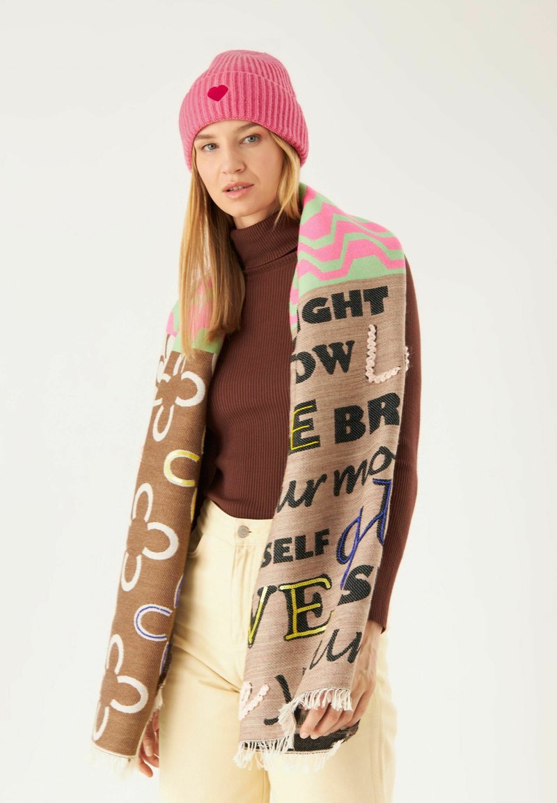 Gebreide roze beanie met een hartdetail; kleurrijke, gepatroonde sjaal met bruin, crème en tekst in zwart, blauw en geel, met franjeranden.