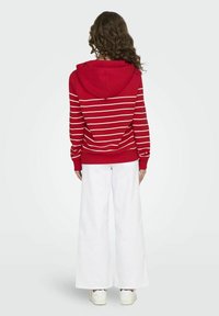 Felpa con cappuccio rossa e strisce orizzontali bianche, con maniche lunghe e un fit rilassato, abbinata a pantaloni bianchi larghi e sneakers.