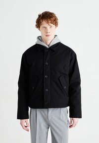 Black jacket with a high collar, front buttons, and side pockets. Worn over a gray hooded sweatshirt. Straight-cut design, contrasting textures.