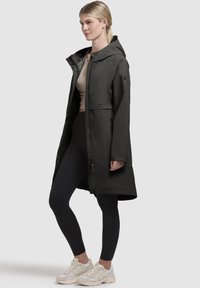 Svart vattentät jacka med huva, tryckknappar och sidofickor. Bärs över en beige topp och svarta leggings, tillsammans med ljusa sneakers.