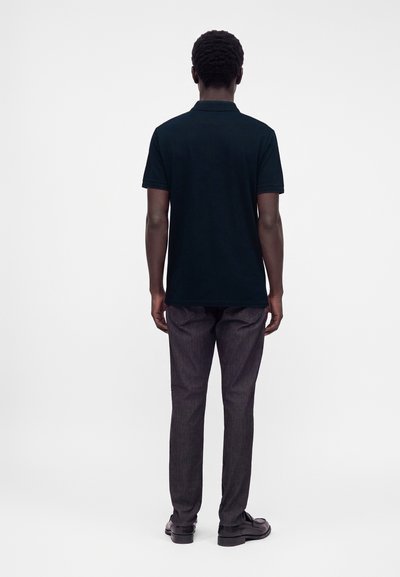 Homme debout de dos, portant un polo noir à manches courtes, un pantalon sombre et des chaussures noires sur fond blanc.