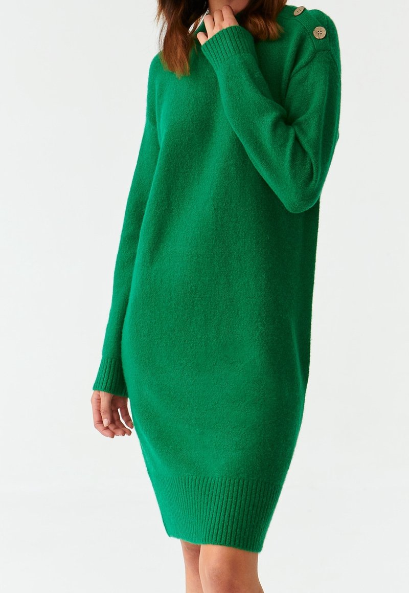 Femme portant une robe-pull verte à manches longues, longueur genou, avec des détails de boutons sur l'épaule, sur fond blanc.