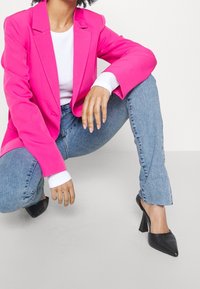 En hetrosa skräddarsydd kavaj över en vit topp, i kombination med ljusblå jeans. Svarta spetsiga klackar fulländar outfiten.