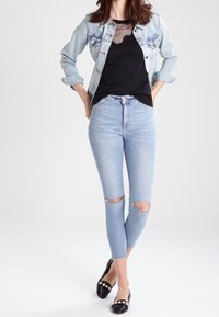 Džínová bunda, černý síťovaný top a světle modré potrhané skinny džíny. Černé baleríny s dekoračními akcenty. Minimalistický styl na světlém pozadí.