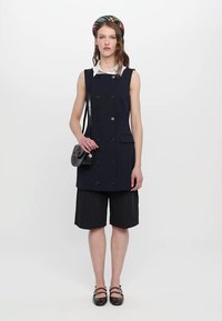 Rochie fără mâneci, de tip dublu-breasted, în nuanță de marină, cu buzunare, combinată cu pantaloni scurți cu dungi. Modelul ține o geantă neagră tip crossbody și poartă o bentiță cu model.