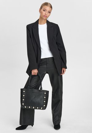 Kvinde iført sort blazer, hvid skjorte og læderbukser holder en sort nittebeslået håndtaske og står mod en ensartet lys baggrund.