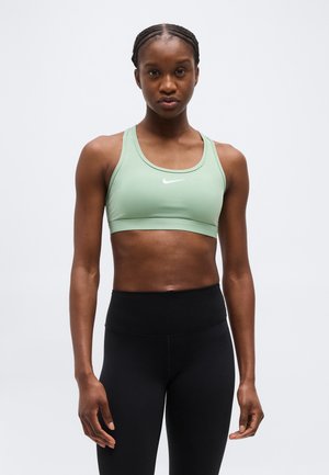 Mujer atlética con trenzas, lleva un sujetador deportivo Nike verde claro y leggings negros de talle alto, posando frente a un fondo liso.