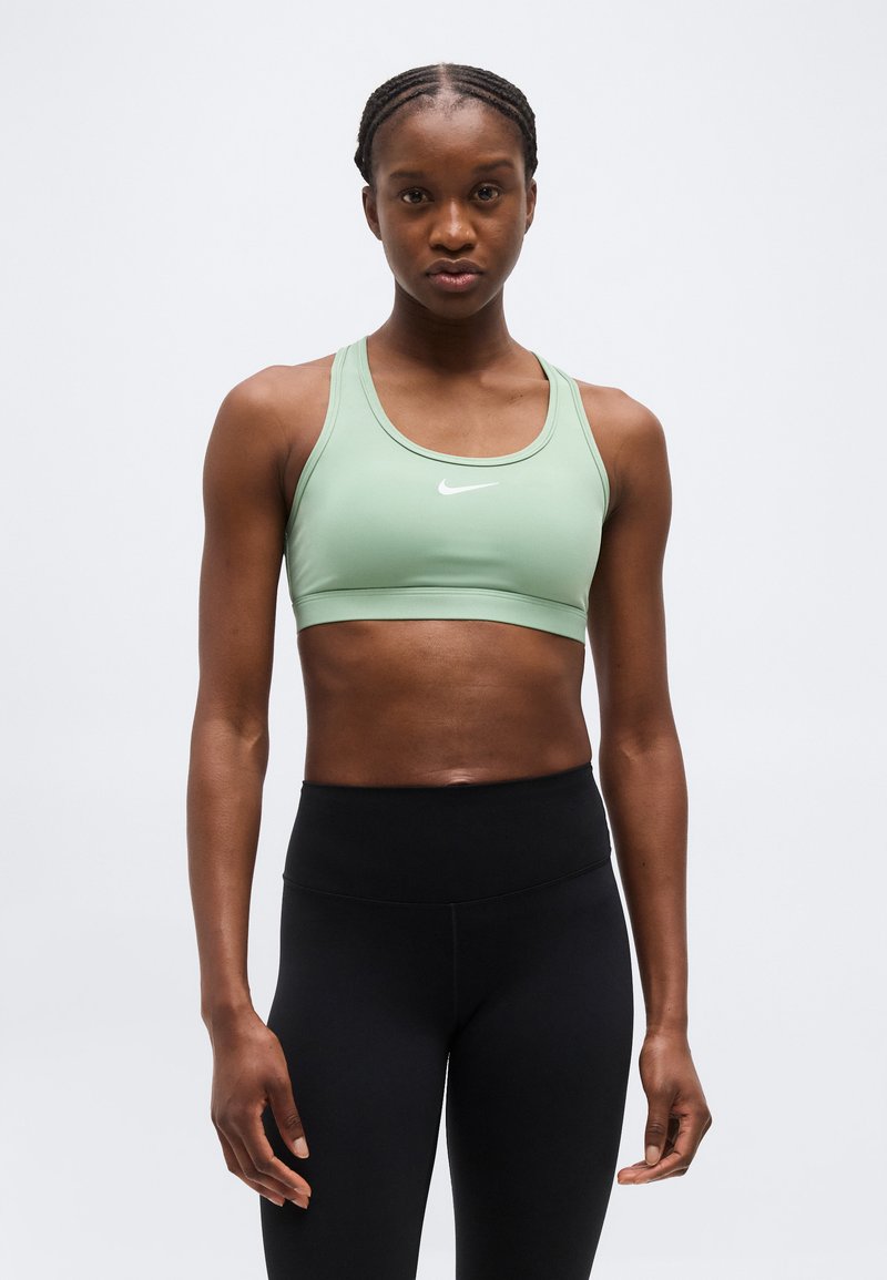 Αθλητική γυναίκα με πλεξούδες, φορώντας ανοιχτό πράσινο αθλητικό σουτιέν Nike και μαύρο κολάν ψηλόμεσο, όρθια μπροστά σε απλό φόντο.