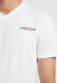 Valge V-kaelusega T-särk, mis on valmistatud puuvillast, millel on väike must ja punane "GUESS JEANS" logo koos "AMERICAN TRADITION" tekstiga selle kohal.