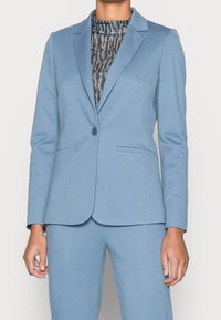 Světle modrý blazer v přizpůsobeném designu, vyrobený z hladké tkaniny s jedním knoflíkem a dvěma předními kapsami, doplněný vzorovaným topem.