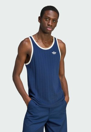 Junger Mann trägt ein blaues Adidas Tanktop mit weißem Rand und passende blaue Hose, Hände in den Taschen, steht vor einem schlichten Hintergrund.