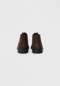 Botas de tobillo de ante marrón con suela de goma negra texturizada, que cuentan con paneles laterales elásticos para facilitar el calzado. Diseño simple y minimalista.