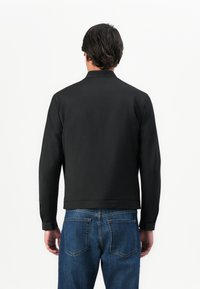 Uomo vestito con una giacca nera a maniche lunghe e jeans blu, in piedi con la schiena rivolta verso uno sfondo bianco uniforme.