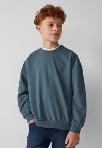 Blauwe katoenen sweatshirt met een ronde hals, een ontspannen pasvorm en geribbelde manchetten. Draagt over een wit shirt, gecombineerd met donkere denim.