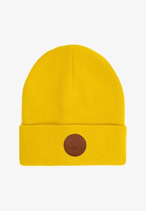 Żółta wełniana czapka z okrągłą skórzaną naszywką, połączona z pasującą żółtą kurtką i białą koszulką, a także ciemnoniebieskimi dżinsami. Długie, kręcone włosy.