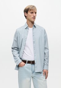 Ανοιχτό μπλε ριγέ πουκάμισο με κουμπιά, μακριά μανίκια, από μαλακό βαμβακερό ύφασμα. Φορεμένο πάνω από ένα λευκό T-shirt και συνδυασμένο με ανοιχτόχρωμο τζιν και καφέ ζώνη.