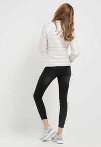 Kvinna med långt hår som bär en vit quiltad jacka, svarta tajta jeans och vita sneakers, stående mot en enfärgad bakgrund.