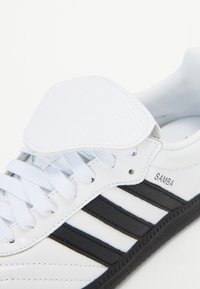 Hvit lær sneaker med tre svarte striper, hvite lisser og en polstret tunge. "Samba"-logo trykt på siden. Gummisåle.